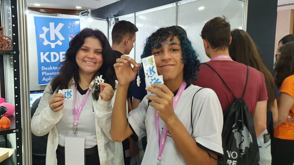 Two happy attendees, a girl and a boy, hold up KDE stickers and keychains.