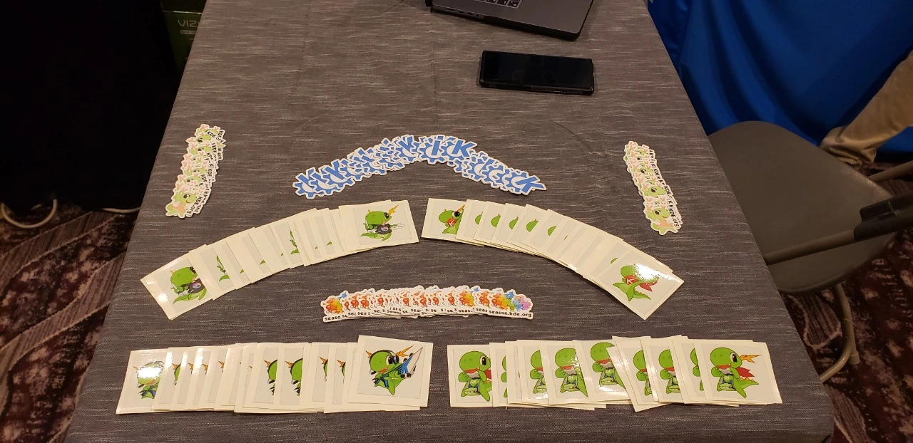 Stickers of Katie, Konqi, and the KDE logo ont the KDE booth's table'.