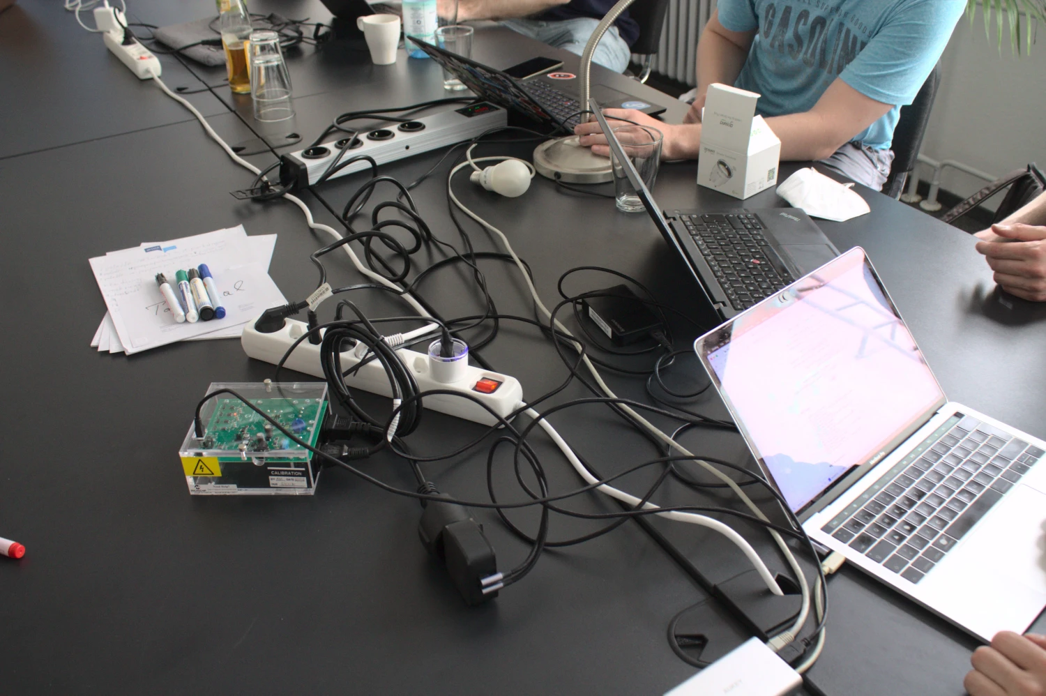 An electronic device in a transparent perspex box sits on a large, black table. The device (GUDE) has several cables connected to it, one going to a special power outlet. KDE Eco team members sit around the table running tests on their laptops.